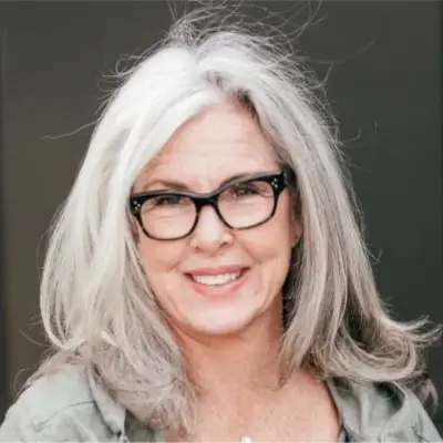A person with long gray and white hair, wearing black-framed glasses and a green top, smiles while looking directly at the camera.