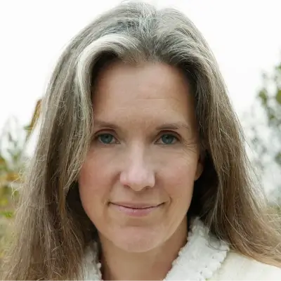 A close-up of a person with long light brown hair featuring a prominent white streak in the front. They have light-colored eyes and are wearing a white textured garment.