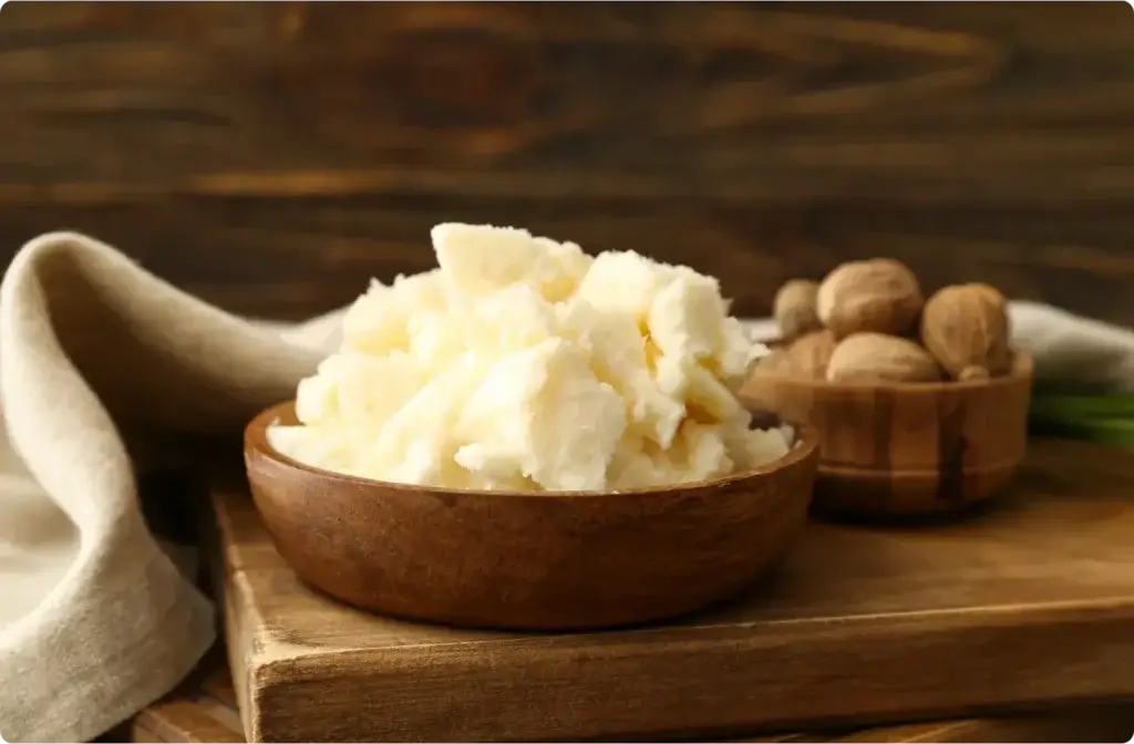 A rustic wooden bowl filled with white, solid shea butter sits beside a smaller wooden container holding whole shea nuts. Both are placed on a wooden surface with a beige cloth in the background, creating a natural, earthy aesthetic.