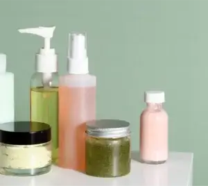 Assorted skincare products arranged on a white surface against a light green background. Items include various pump bottles, spray bottles, and jars containing liquids and creams in shades of green, yellow, pink, and cream, suggesting a range of cosmetic or personal care formulations.
