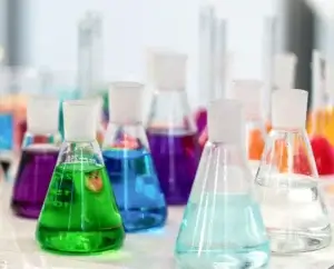Collection of Erlenmeyer flasks filled with brightly colored liquids—green, blue, purple, orange, red, and clear—arranged on a white surface.
