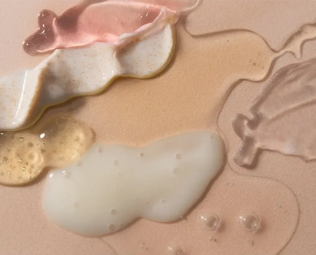 Various skincare product textures displayed on a smooth beige surface, including pearlescent pink gel, white cream with exfoliating particles, translucent golden gel, milky white lotion, and clear serum. Each smear or droplet highlights differences in consistency and formulation.