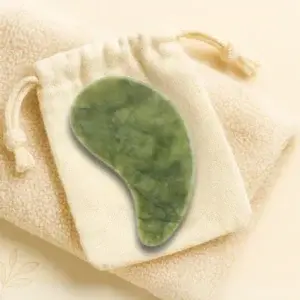 Green jade gua sha tool resting on a beige drawstring pouch atop a soft, textured towel. The tool has a smooth, curved shape designed for facial massage. The background is light cream, creating a calm and soothing aesthetic.
