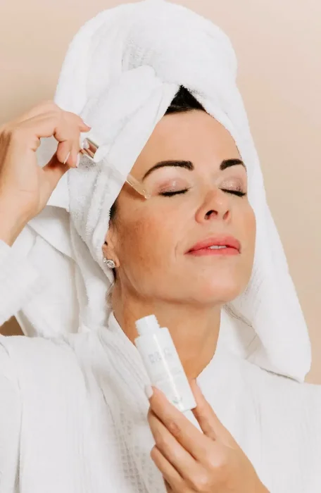 A person wearing a white robe and a white towel wrapped around their head applies skincare serum near the eye using a dropper.